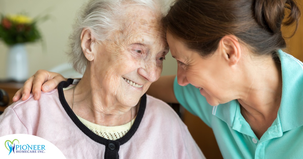 A caregiver lovingly embraces a senior woman while they both smile, representing the kind of warm disposition that can help when dealing with dementia-related repetition.